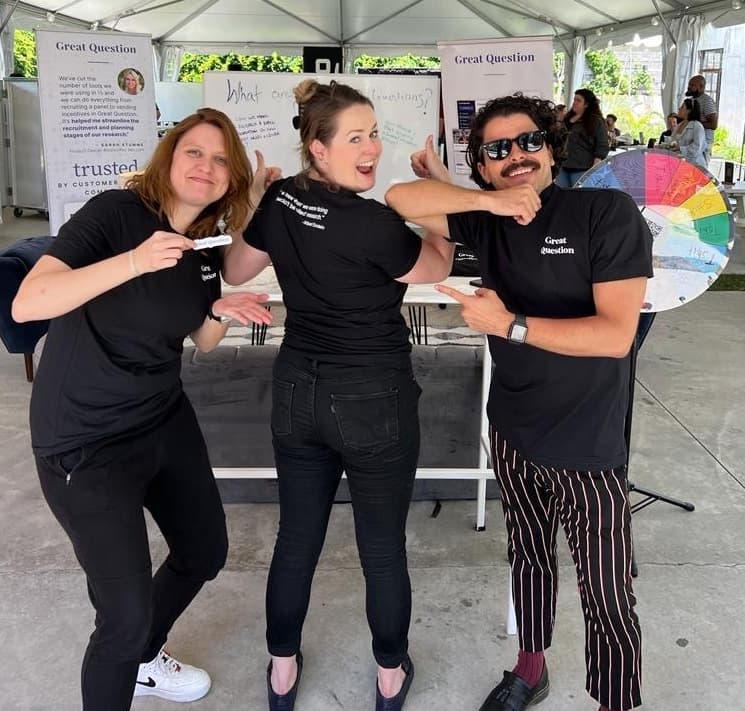 Three Great Question team members posing at the company's booth at UXRConf 2022, pointing at their branded t-shirts beside a prize wheel and a whiteboard of visitor questions.
