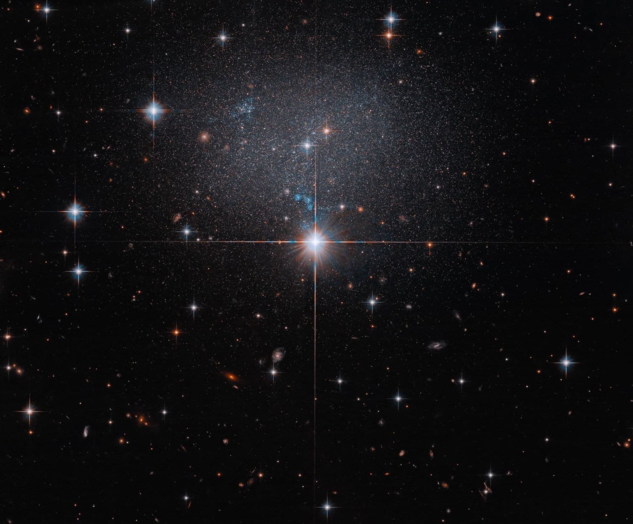 The faint, low-surface-brightness dwarf irregular galaxy ESO 490-017, a diffuse swarm of stars behind bright foreground stars, set against a field of distant reddish galaxies.