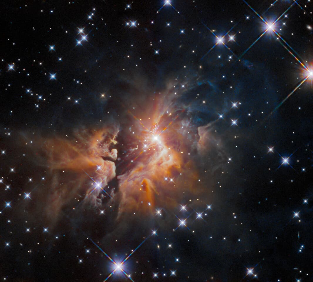 The bright young stellar object IRAS 05506+2414 cloaked in thick gas and dust, with a fan-shaped spray of glowing material streaming outward across the frame.