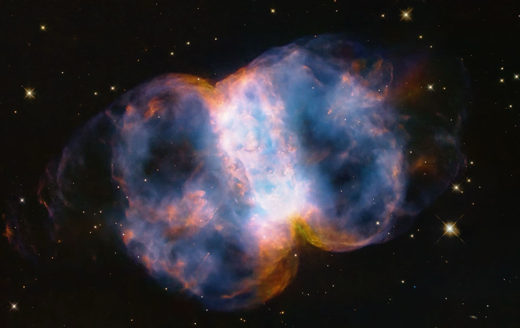 Hubble image of the Little Dumbbell Nebula (M76), a glowing two-lobed planetary nebula in blue and orange against a dark star field.
