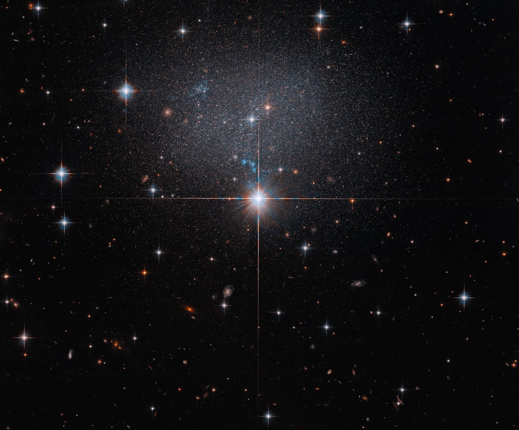 The faint, low-surface-brightness dwarf irregular galaxy ESO 490-017, a diffuse swarm of stars behind bright foreground stars, set against a field of distant reddish galaxies.