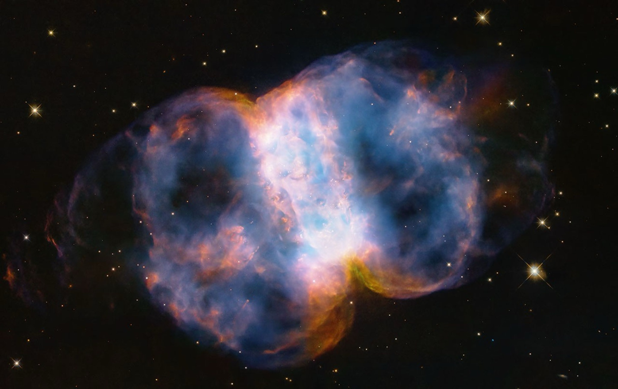 Hubble image of the Little Dumbbell Nebula (M76), a glowing two-lobed planetary nebula in blue and orange against a dark star field.